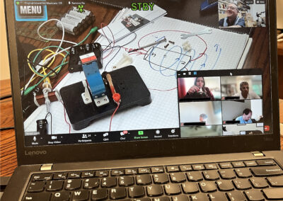 Laptop screen showing a video call; main window shows a cluttered electronics workspace with wires, a notebook, and small components; a person smiles in the top-right video tile.