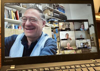 Close-up of a smiling man wearing glasses during a video call on a laptop, with other participants in small tiles on the right and a cluttered office background.